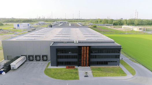 Nowoczesny magazyn i biurowiec przemysłowy w Polsce, widok z lotu ptaka przedstawiający dużą halę produkcyjną z dokami załadunkowymi, samochodem dostawczym oraz otaczającymi zielonymi polami.